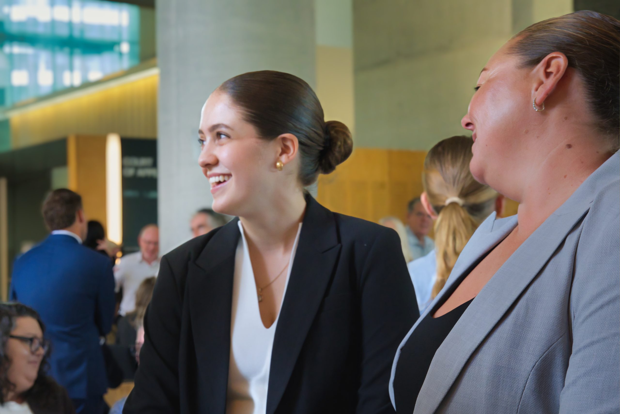 Mikaela Egan smiles and looks to her right while speaking with colleagues inside the Supreme Court of Queensland following her admission ceremony, wearing a black blazer over a white top with her hair neatly styled in a bun.